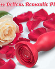 Red rose vibrating plug resting among rose petals with its wide flower base, underside power button and two magnetic charging contacts facing forward