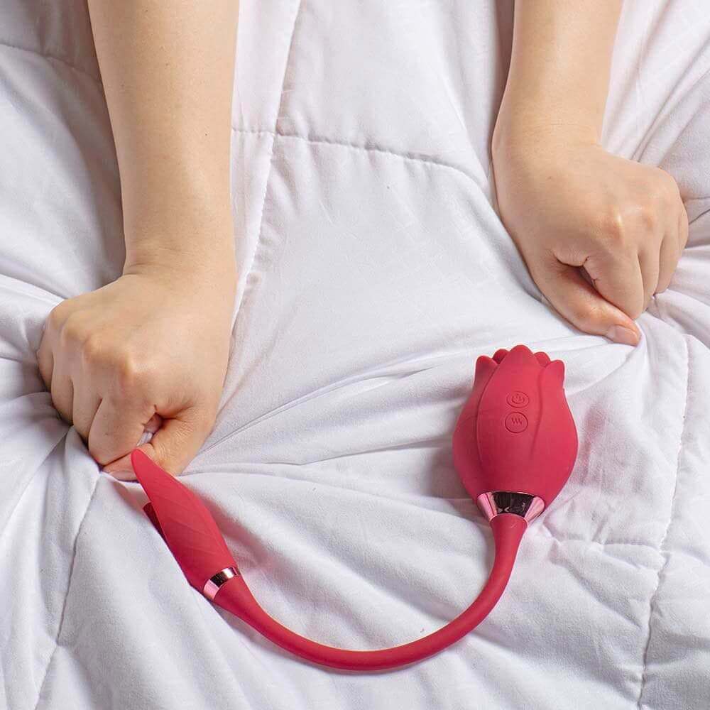 Pink Mermaid rose suction vibrator on white bedding with a person holding the petal-shaped connected egg, showing the flexible U-shaped cord and two body buttons