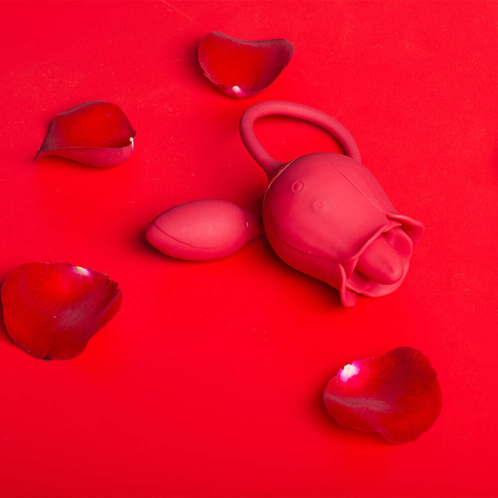 Red rose tongue vibrator with two side buttons and a connected egg arranged on a red background with loose rose petals