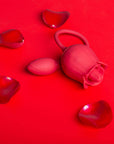 Red rose tongue vibrator with two side buttons and a connected egg arranged on a red background with loose rose petals