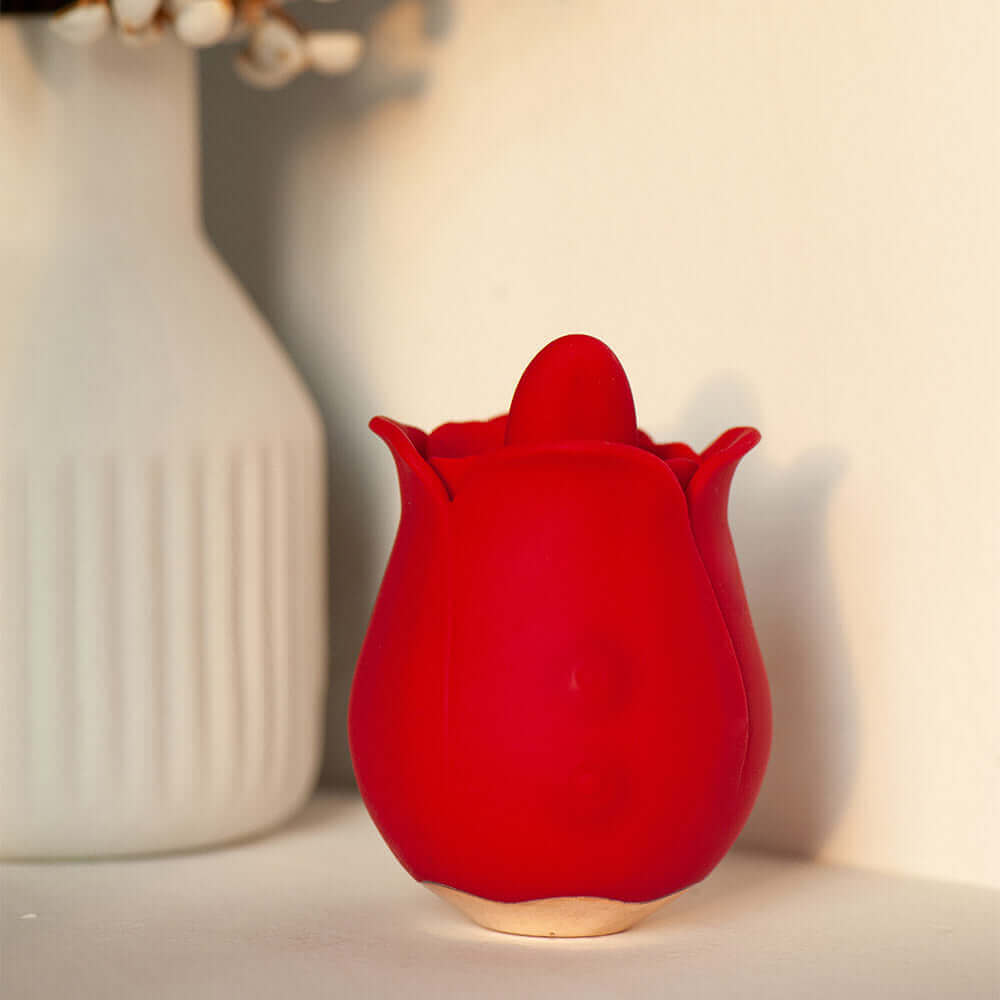 Red compact rose tongue vibrator with a raised central tongue and gold-colored base beside a white vase
